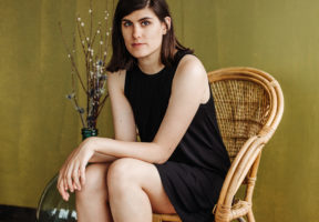 A white woman with long dark hair and dark bangs sits in a high-backed wicker chair against a beige-green wall. She wears a black sheath dress with open arms.