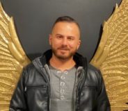 A Syrian-Canadian man with short brown hair and a small goatee. He wears a grey t-shirt and a dark leather jacket and stands against a wall with golden wings that stretch out from his shoulders.