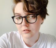 A non-binary individual with short tousled brown hair and dark-rimmed glasses. They wear a white t-shirt and are sitting against a cream-coloured wall.