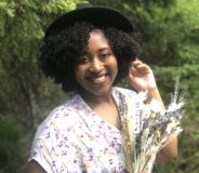 A young Black woman wears a white floral print dress and stands against a leafy green background.