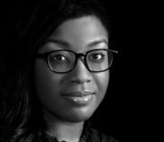 A black-and-white photo of a young Black woman against a black backdrop. She wears dark-rimmed glasses and is smiling slightly.