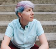 A white trans man with shourt pale blue and pink hair sits on a flight of grey concrete steps, looking off to the side. They wear a light blue button-down t-shirt.