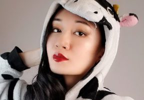 An Asian woman with dark red lipstick stands against a beige background. She is wearing a cow snuggly suit.
