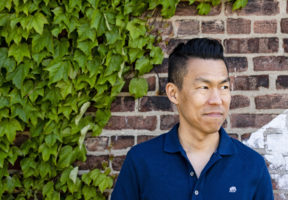 Photo of author Jack Wang--a middle-aged Chinese-Canadian man with short dark hair, wearing a dark blue button-up t-shirt. He stands against a brick wall and looks off to his left.