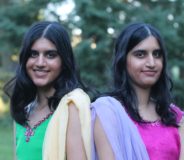 Photo of two young identical Indo-Canadian women standing back-to-back with one another. They both have long dark hair, dark eyes, and are smiling. The sister on the left wears green and yellow and the sister on the right wears purple and pink.