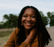 Photo of author Bethaney Wilkinson--a Black woman with long dark hair, sitting down in a lush green field