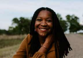 Photo of author Bethaney Wilkinson--a Black woman with long dark hair, sitting down in a lush green field