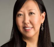 A middle-aged Asian-Canadian woman with long dark hair. She wears dangling earrings and a black button-up shirt and is smiling faintly at the camera.