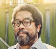 A middle-aged Black man with short dark hair and a salt-and-pepper beard. He wears glasses and a light grey button-up shirt and stands in front of a wooden fence.