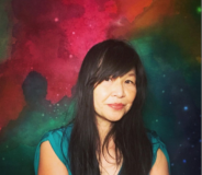 A middle-aged Asian-Canadian woman with long dark hair sits against a multi-coloured abstract painting.