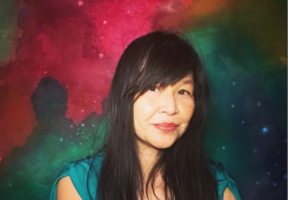A middle-aged Asian-Canadian woman with long dark hair sits against a multi-coloured abstract painting.