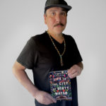 A middle-aged Indigenous man with short dark hair and a mustache. He wears a dark t-shirt and holds a book, titled LIFE IN THE CITY OF DIRTY WATER. He also wears a dark baseball cap.