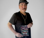A middle-aged Indigenous man with short dark hair and a mustache. He wears a dark t-shirt and holds a book, titled LIFE IN THE CITY OF DIRTY WATER. He also wears a dark baseball cap.
