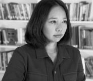 Black and white photo of a young Tibetan-Canadian woman with shoulder-length dark hair, wearing a dark button-up shirt and standing against a white shelf filled with books.