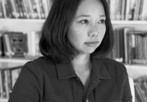 Black and white photo of a young Tibetan-Canadian woman with shoulder-length dark hair, wearing a dark button-up shirt and standing against a white shelf filled with books.