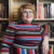 A fat white woman in her late 30s stands in front of a full bookshelf. The photo shows her from the waist up; she's wearing a fitted multi-coloured striped dress with a mockneck and long sleeves. She has chin-length curled golden brown hair, wears square light blue glasses, orange lipstick and green pompom earrings.