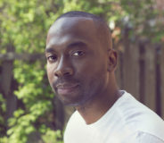 A young Black man with a shaved head and a small goatee. He wears a white t-shirt and is sitting outside. In the background is a lush green tree.