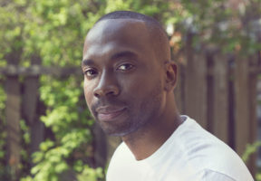 A young Black man with a shaved head and a small goatee. He wears a white t-shirt and is sitting outside. In the background is a lush green tree.