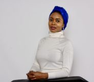 A black woman wearing a purple headscarf and a cream turtleneck, sitting against a white wall.
