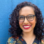 A middle-aged Indian-Canadian woman with curly dark hair and glasses. She stands against a blue wall and wears a multi-coloured blouse and red lipstick. She is smiling widely.