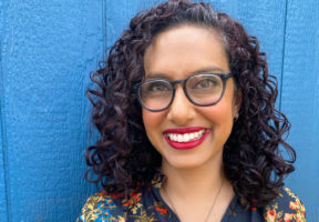 A middle-aged Indian-Canadian woman with curly dark hair and glasses. She stands against a blue wall and wears a multi-coloured blouse and red lipstick. She is smiling widely.