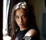 An Asian transwoman with long dark hair, standing by a window and looking directly at the camera with a serious, intense look on her face.