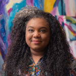 A Black woman with long curly dark hair smiles softly. She stands against a multi-colouted painting in the background.