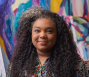 A Black woman with long curly dark hair smiles softly. She stands against a multi-colouted painting in the background.