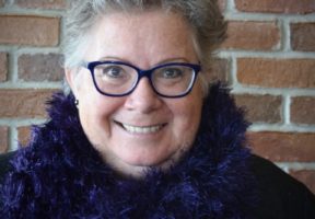 Photo of a senior white woman with short grey hair, glasses, and wearing a purple sweater against a brick background.