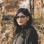 A photo of a young Indo-Canadian woman with long dark hair and dark-framed glasses, standing against an autumn-leaf nature background.