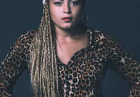 A young mixed-race woman with long brown-blonde dreads caught up in a white scarf. She stands against a dark background and faces the camera with her hands on her hips.