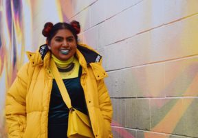 A young Indian-Canadian woman wearing a bright yellow jacket, standing next to an ivory brick wall.