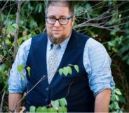 A photo of author S. Bear Bergman--a large trans man with a beard, glasses and short dark hair, wearing a dark blue vest over a light blue button-up shirt-