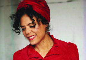 A photo of a middle-aged biracial woman with curly dark hair held in a red scarf and wearing a red shirt.