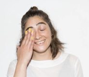 A woman of colour with shjoulder-length brown hair wears a long-sleeved white top. SHe stands against a white background and is laughing, with her right hand half covering her face.