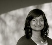 A black and white photo of an Indian-Canadian woman with long dark hair and bangs. She wears a dark top over a white tank top and is smiling softly.