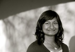 A black and white photo of an Indian-Canadian woman with long dark hair and bangs. She wears a dark top over a white tank top and is smiling softly.